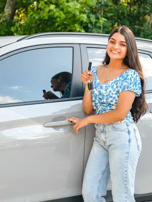 Smiling young woman holding car keys while standing next to a silver SUV with trees in background
