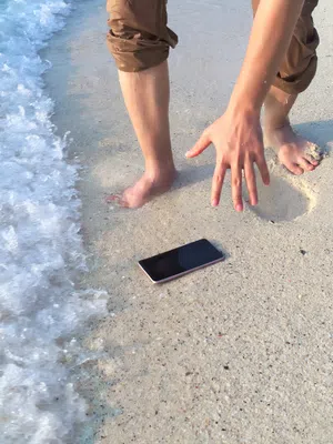 Person reaching to rescue a smartphone from wet sand near incoming ocean waves on beach shore.