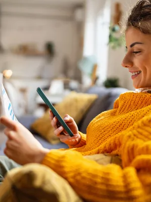 Smiling woman in a mustard sweater using smartphone while holding paper on a cozy living room couch.