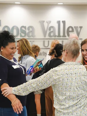 Smiling diverse group warmly greeting and embracing at Coosa Valley Community event in indoor setting.