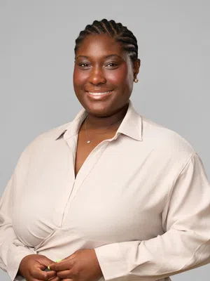 Smiling Black woman with braided hair wearing a beige shirt and small necklace against a plain gray background
