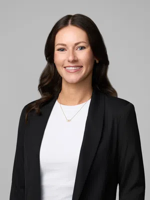 Professional woman with long brown hair smiling, wearing black blazer, white shirt, and gold necklace on gray background