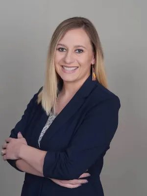 Smiling woman with blonde hair wearing a navy blazer and colorful earrings posing with arms crossed