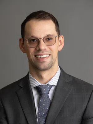 Smiling man in glasses wearing a gray suit and patterned tie standing with arms crossed against gray background