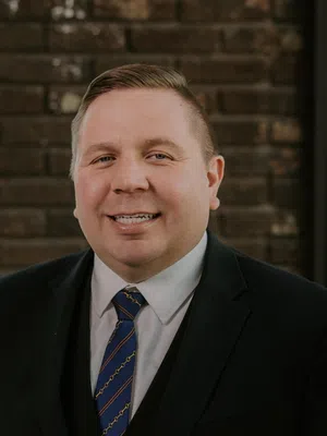 Smiling man in black suit and blue striped tie standing in front of a brick wall near a window.