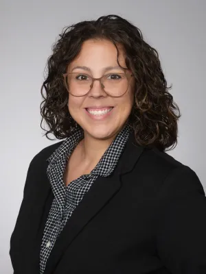 Smiling woman with curly hair wearing glasses, a black blazer, and a checkered shirt against a gray background.