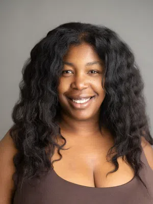 Smiling woman with long curly black hair wearing a brown sleeveless top on gray background.