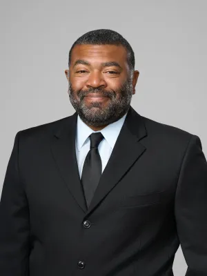 Confident man in black suit and tie smiling against a plain gray background, professional studio portrait.