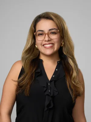 Smiling woman with long highlighted hair wearing glasses and a black ruffled sleeveless top against gray background