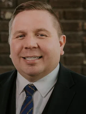 Smiling man wearing a black suit and blue striped tie standing in front of a brick wall indoors.