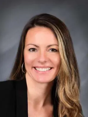 Professional portrait of a smiling woman with long blonde hair wearing a black blazer and hoop earrings.