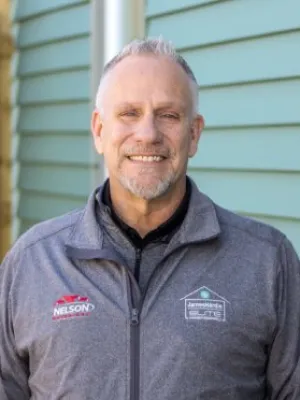 Smiling man with gray hair wearing a gray jacket with Nelson and James Hardie logos in front of green siding.