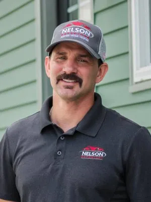 Man wearing a Nelson Roofing hat and polo shirt standing outside a green house siding background.
