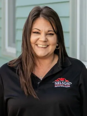 Smiling woman with long brown hair wearing a black Nelson branded polo shirt outdoors near a house.