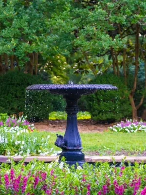 Black birdbath fountain surrounded by blooming purple and white flowers in a lush green garden.