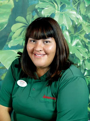 Smiling woman in green Armstrong Garden Center shirt with dark hair standing in front of large leafy plants.