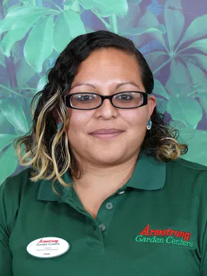 Woman wearing green Armstrong Garden Centers polo and name tag stands against leafy green backdrop