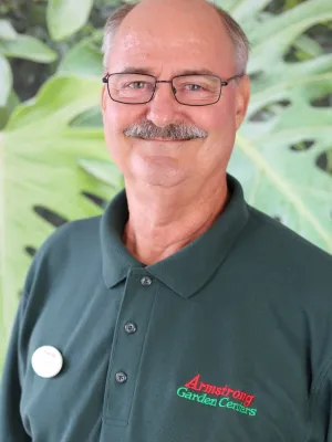 Smiling man with glasses wearing a dark green Armstrong Garden Centers polo outdoors with large green leaves in background
