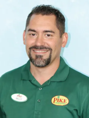 Man wearing a green Pike Nurseries shirt with name tag Billy smiling against a light blue background