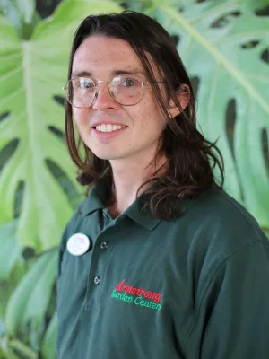Young woman wearing glasses and Armstrong Nature Center polo standing in front of leafy green plants smiling.