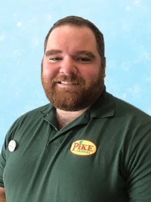 Smiling man with beard wearing a dark green Pike Nurseries polo shirt against a light blue background