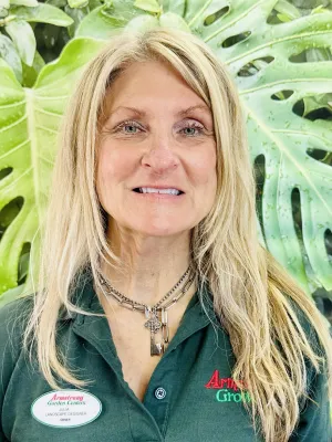 Smiling woman with blonde hair wearing Armstrong Garden Centers shirt and key necklace against leafy green background