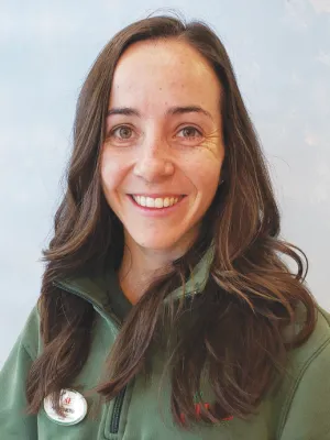 Smiling young woman with long brown hair wearing a green jacket and a name tag against a light background