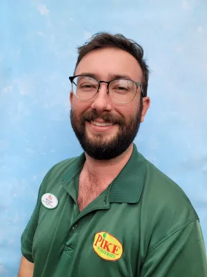 Smiling man with glasses and beard wearing green Pike Nurseries polo and name tag against blue background