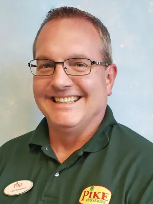 Smiling man wearing glasses and green Pike Nurseries polo shirt with name tag on light blue background