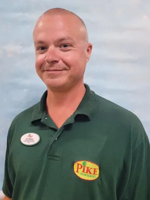 Smiling man wearing dark green Pike Nurseries polo and name tag on light blue background