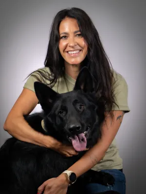 Smiling woman with long dark hair hugging a happy black German Shepherd dog with tongue out against gray background
