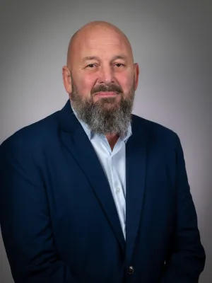 Portrait of a bald man with a thick beard wearing a navy blue suit and white shirt on a gray background