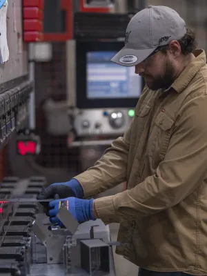 Worker in a cap and gloves measures a metal piece using calipers in a factory setting with machinery.
