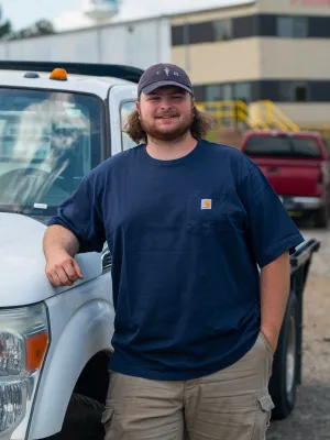 Man in navy shirt and cap leaning on white truck in a parking lot with other vehicles in background
