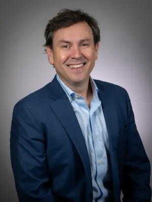 Smiling man in blue suit jacket and light blue shirt against plain gray background.