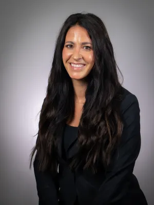 Smiling woman with long dark hair wearing a black blazer against a plain gray background.