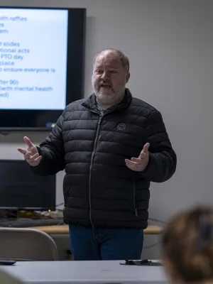 Man giving a presentation in a classroom with a screen displaying employee benefits and programs