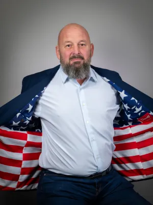 Bearded man in white shirt and jeans reveals American flag patterned jacket lining against a gray background.