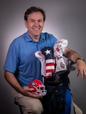 Man in blue polo holding a red football helmet beside a golf bag with patriotic and embroidered club covers.