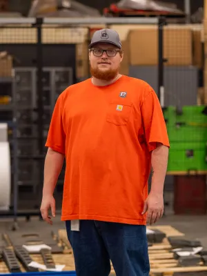 Man wearing orange shirt and cap stands in industrial warehouse surrounded by wooden pallets and metal racks