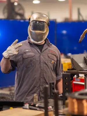 Welder wearing protective gear showing peace sign in industrial workshop with welding equipment