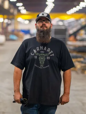 Bearded man in a Carhartt t-shirt and cap stands in a large industrial warehouse holding a tool.