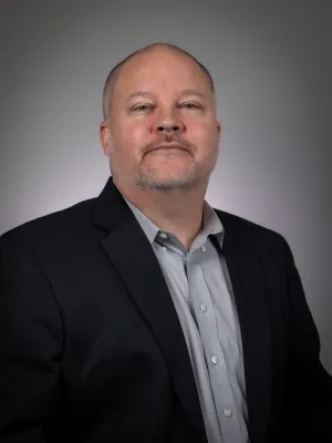 Professional portrait of a bald man with a beard wearing a black suit jacket and gray shirt against a gray background