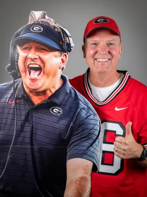 Smiling man in red Georgia Bulldogs jersey and hat standing next to cardboard cutout of enthusiastic football coach.