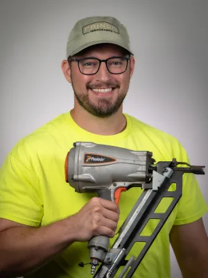 Smiling man in yellow shirt and cap holding a Paslode nail gun on a gray background.