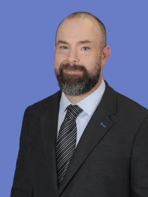 Smiling bald man with a beard wearing a black suit, light blue shirt, and striped tie against a blue background.