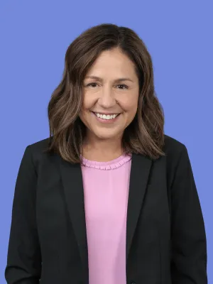 Smiling woman with shoulder-length brown hair wearing a black blazer and pink top against a blue background