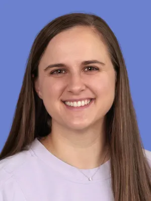 Smiling woman with long brown hair wearing a light purple shirt and a small pendant necklace against blue background