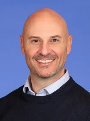 Smiling bald man with light stubble wearing a navy sweater over a collared shirt against a blue background