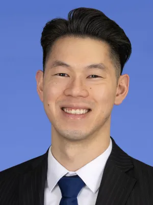 Smiling young Asian man wearing a black suit, white shirt, and blue tie against a blue background